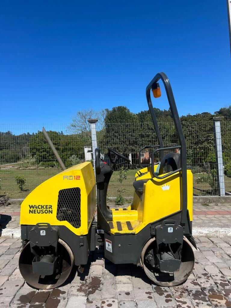 Rolo compactador Wacker Neuson RD12 estacionado sobre pavimento de blocos, sob céu azul.